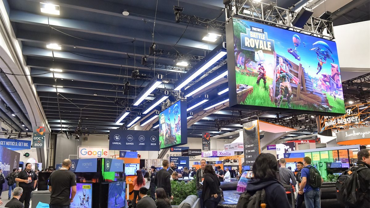Telão com a arte do Fortnite Battle Royale pendurado sobre o estande da Epic Games numa feira de games, com visitantes jogando embaixo