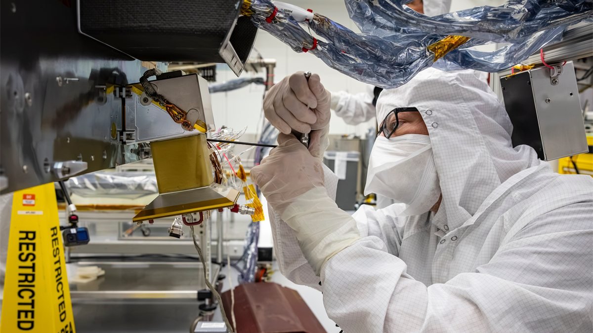 Técnico com roupa de sala limpa, máscara e luvas instala uma das câmeras de navegação do Dragonfly, uma peça dourada sob a estrutura do drone, ao lado de cabos envoltos em plástico