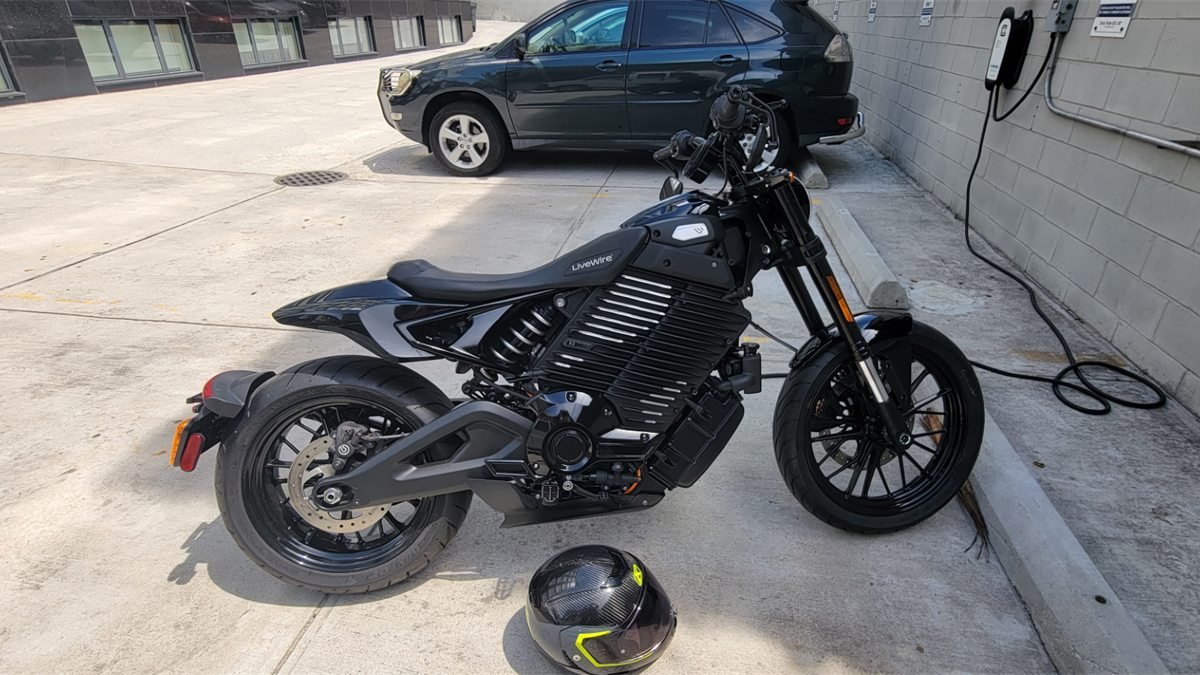 Moto elétrica preta da LiveWire estacionada ao lado de um carregador de parede, com um capacete no chão