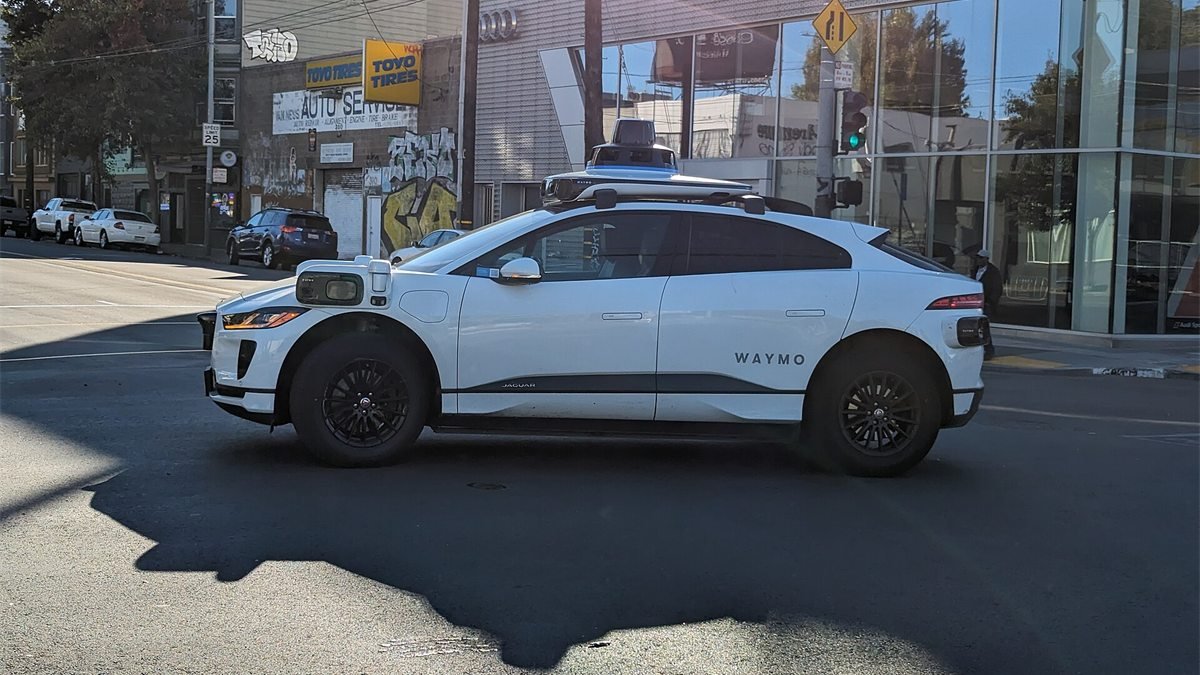 Carro branco da Waymo com sensores no teto e nos para-lamas atravessa um cruzamento em San Francisco, num dia de sol