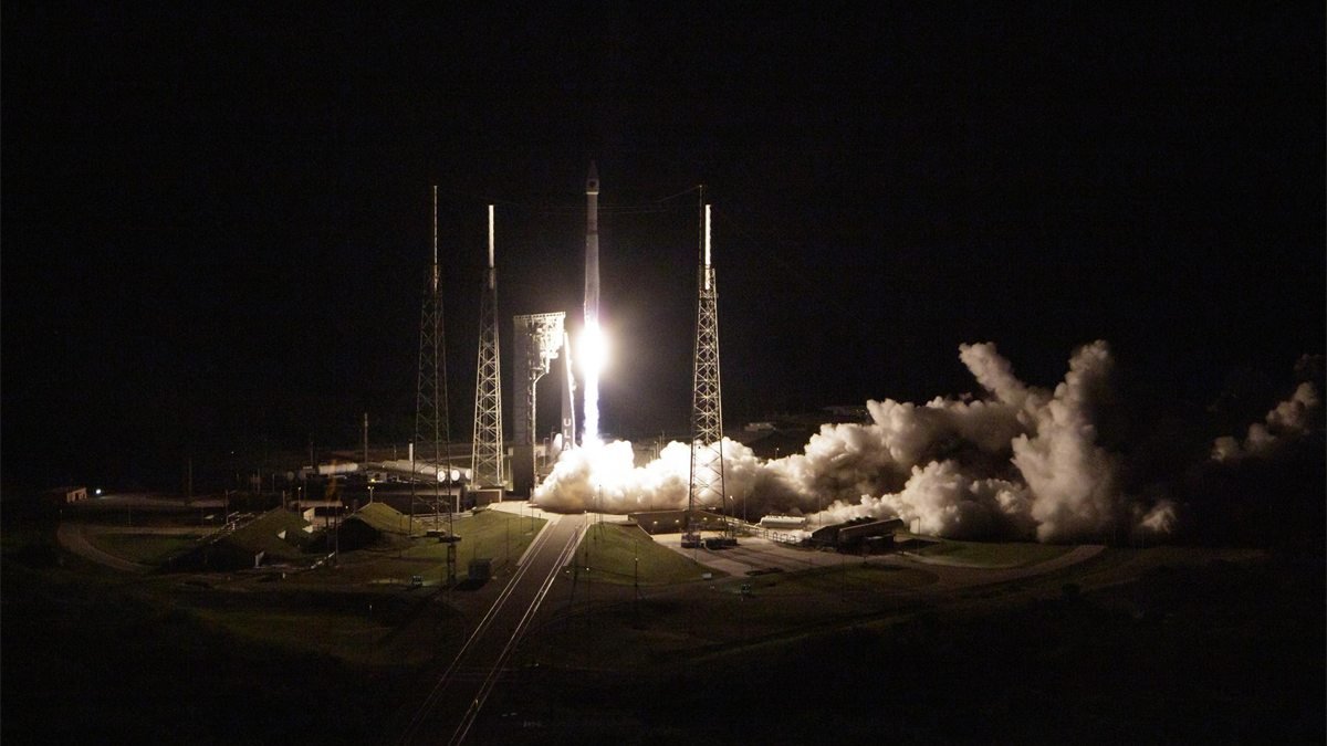 Foguete Atlas V decola à noite da plataforma de lançamento, entre torres metálicas, com uma grande nuvem de vapor branco à direita