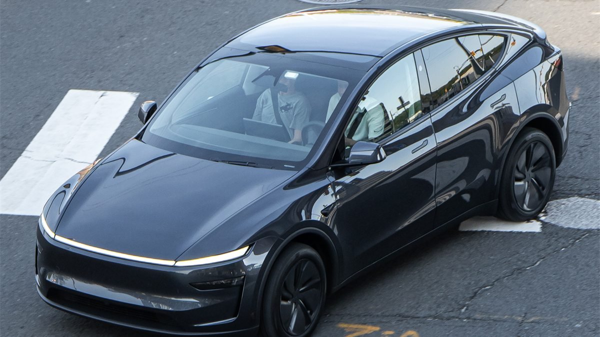 Tesla Model Y cinza-escuro visto de cima atravessa uma faixa de pedestres, com duas pessoas nos bancos da frente